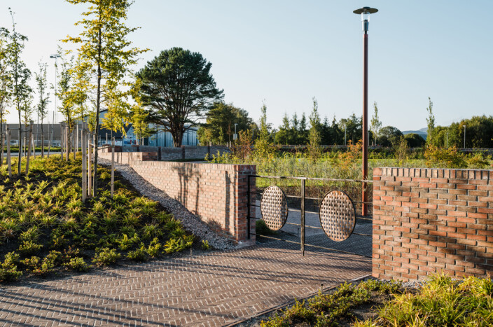 Inngang fra sørvest via buffersone med bøketrær og mur. Porten er utsmykket med flettet kobber. I bakgrunnen ses et flott furutre som ble bevart. Inngang fra sørvest via buffersone med bøketrær og mur. Porten er utsmykket med flettet kobber. I bakgrunnen ses et flott furutre som ble bevart.